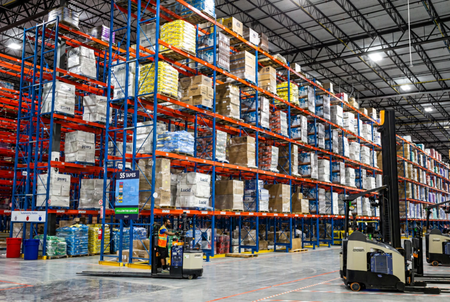 Stocked warehouse pallet racks with reach trucks, a lift operator and marked floor lanes